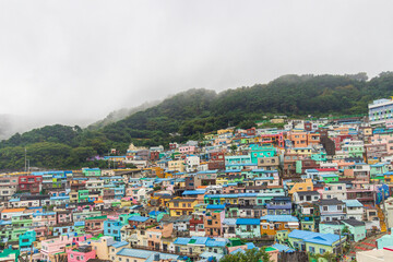 Dense rows of colorful homes cover a forested hillside beneath drifting low fog and an overcast sky. © Saigonese