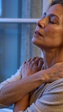Vertical video: Adjusting senior woman easing shoulder tension beside window, with sweater and ring