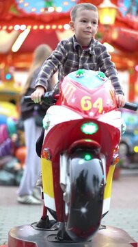 Happy little boy laughs on a colorful carnival motorcycle ride at night amusement park, holding handlebars under bright festive lights.