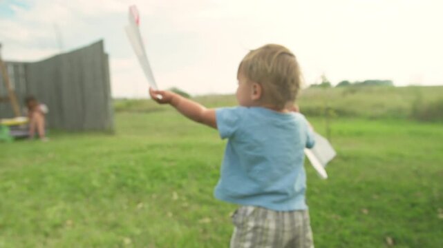Toddler walking through backyard holding paper airplanes near swing set