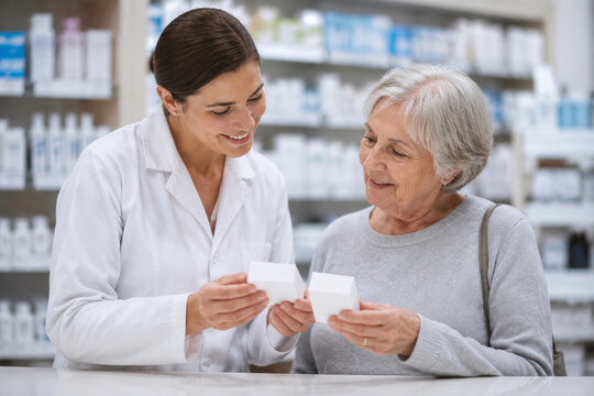 Friendly female pharmacist showing medication packaging to elderly woman inside community pharmacy store with shelves of medical supplies in background.