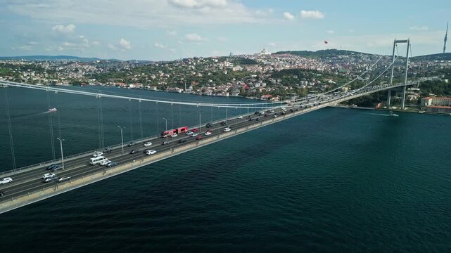Turkey, Istanbul 01 10 2025: Drone video moving backwards from Bosphorus Bridge traffic to a wider view of the bridge with sailing boats and Camlica Mosque on a cloudy day.