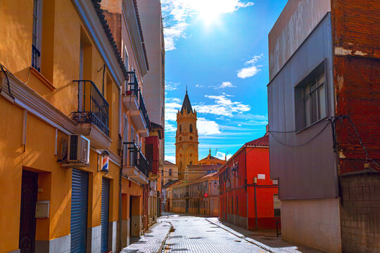 Street in Malaga leads toward the historic Iglesia de San Pablo tower. Bright sunlight illuminates yellow facades and cobblestones, capturing a quiet afternoon in Andalusia from Spain
