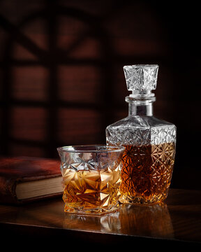 Vaso y licorera de cristal tallado con whisky o bourbon junto a libro antiguo sobre mesa de madera barnizada