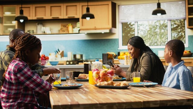 African American mother appears detached from her family at the breakfast table while distracted by phone use, showing divided attention, disconnection, and an unfocused family moment at home.