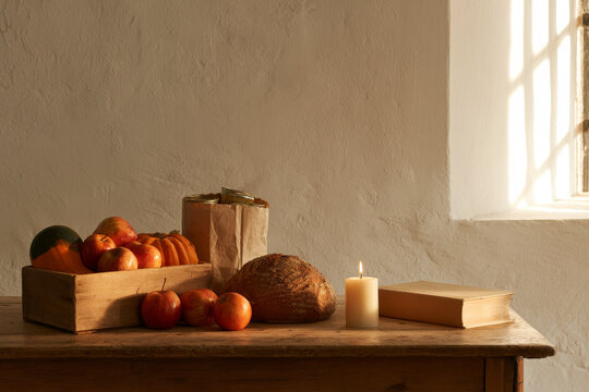 A plain wooden table set for harvest thanksgiving holds squash and apples, a round loaf, a lit candle and tinned food bagged for a food bank, in soft October light.