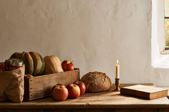 A plain wooden table set for harvest thanksgiving holds squash and apples, a round loaf, a lit candle and tinned food bagged for a food bank, in soft October light.