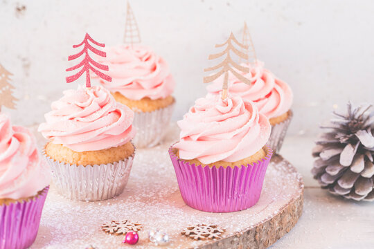 Five vanilla sponge Cupcakes with pink buttercream icing decorated with Christmas trees on a table with a pinecone 
