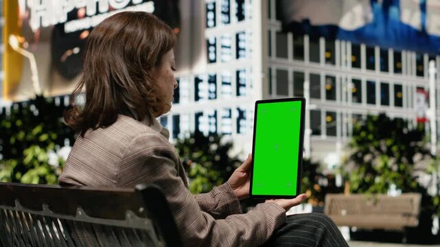 Businesswoman uses a green screen tablet on a park bench to catch up on online news, browsing content on the mockup device during personal time outdoors.