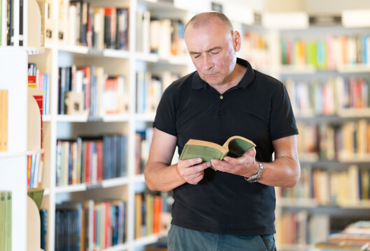 Attentive middle-aged man choosing books near high wooden shelves in bookstore aisle