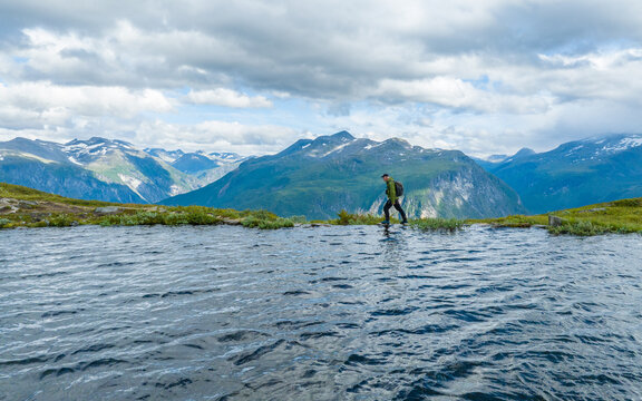 Aerial view of a hiker with a backpack walking along the edge of a mountain lake near Vardebu with rugged peaks and a cloudy sky in Valldal, More og Romsdal, Norway.