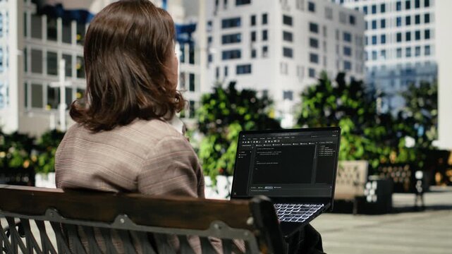 Female programmer writes code on a laptop from a park bench, developing software on the notebook display while working remotely outdoors.