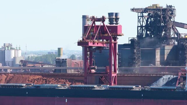 Aerial drone view of a cargo ship loading at a port next to a metallurgical steel plant with smoke emissions. Industrial maritime logistics and environmental impact.