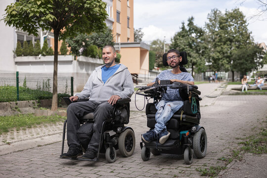 Two powered wheelchair users enjoying a conversation while moving along an accessible sidewalk in an urban environment.