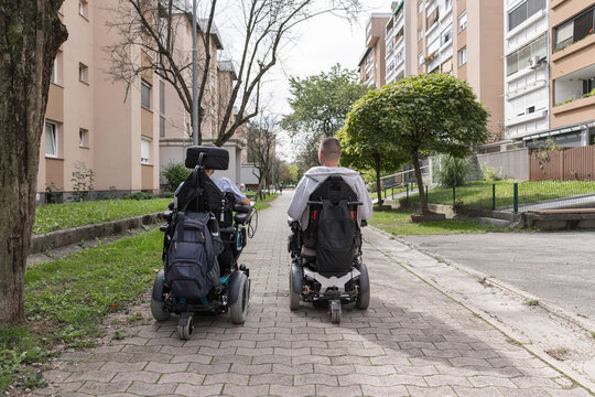 Two men using powered wheelchairs navigating an accessible city walkway, enjoying independent mobility together.
