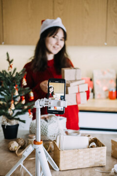 Phone on tripod records woman in Santa hat carrying stack of wrapped parcels in kitchen. Influencer marketing holiday season, small business video promotion, content creator business marketing