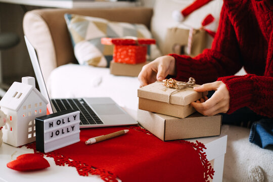 Hands stack kraft boxes beside laptop on table with HOLLY JOLLY sign and ceramic house. Returning unwanted christmas gift, gift exchange policy, post holiday returns