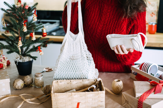Woman holds mesh bag and rolled cotton towel above basket with gift supplies on table. Eco friendly christmas gift set, sustainable home care package christmas, zero waste christmas basket