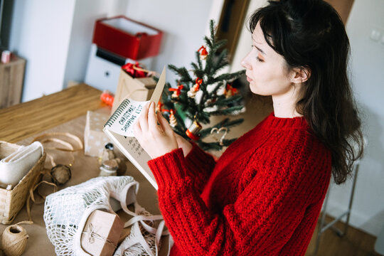Woman turns page of notebook with CHRISTMAS SHOPPING LIST beside wrapped gifts and small tree. Christmas no impulse buying, mindful christmas spending habits, financial discipline holiday season