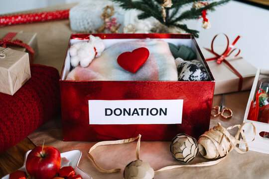 DONATION box filled with knitwear and heart stands on table among wrapped gifts near Christmas tree. Winter clothes donation drive, holiday charity collection box, donate warm clothing christmas