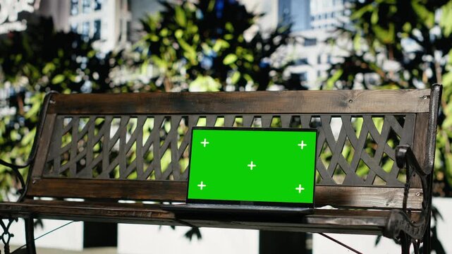 Green screen laptop sits on a wooden bench in an urban park, showing a replaceable chroma key notebook display against metropolitan greenery and downtown buildings in close-up composition.