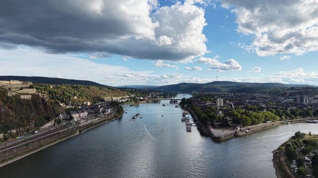Aerial drone view of Koblenz, Germany, showing the confluence of the Rhine and Moselle rivers at Deutsches Eck, with Ehrenbreitstein Fortress and the cityscape under dramatic clouds.