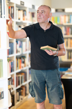 Focused middle-aged guy browsing through literature volumes on shelves in cozy bookshop