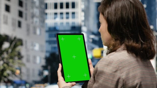 Female professional in business attire holds a green screen tablet outdoors in a business center, using its chroma key touchscreen for content replacement.