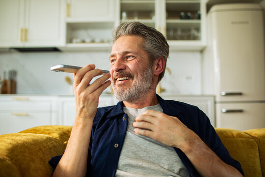 Smiling mature man using smartphone voice assistant at home kitchen