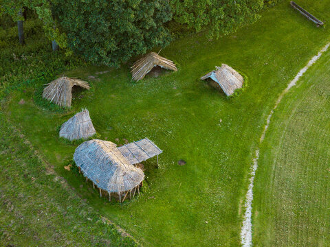 Aerial view of the reconstructed historic thatched-roof dwellings at the Kernave Archaeological Site, set in a lush green meadow with a dirt path in Kernave, Vilnius County, Lithuania.