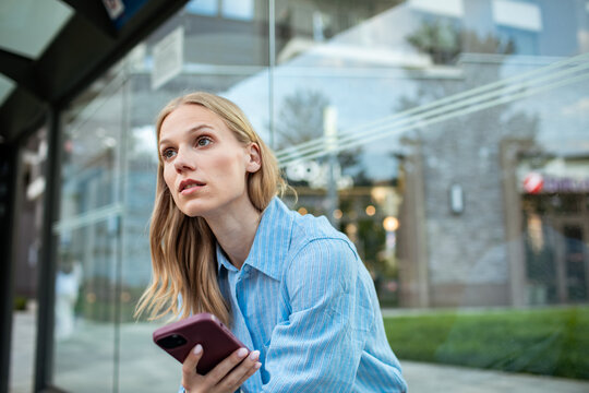 Young woman waiting at city bus stop with smartphone outdoors