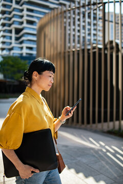 Businesswoman using smartphone while walking downtown with laptop