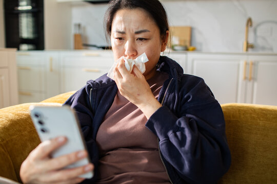 Sick woman looking at smartphone with tissue on living room sofa