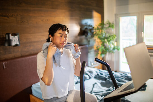 Woman on exercise bike with laptop in home bedroom