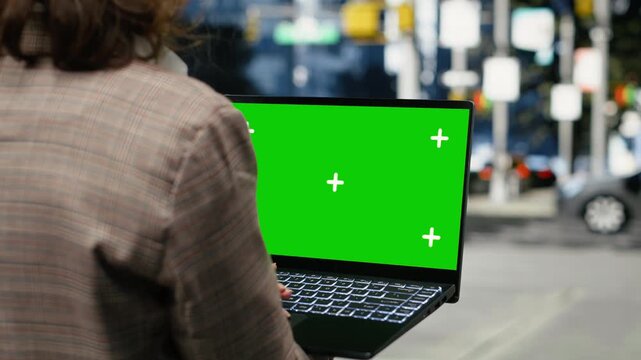 Businesswoman holds a green screen laptop on a city street outdoors, using a chroma key notebook in the downtown center with metropolitan buildings and office architecture nearby in close-up.