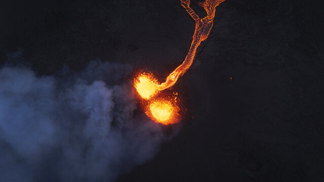Aerial view of glowing lava erupting from an active volcano with thick smoke rising over the dark volcanic landscape in Reykjavik, Reykjavikurborg, Iceland.