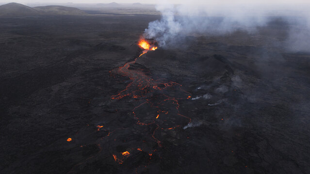 Aerial view of an active volcano erupting with glowing lava flowing across the dark volcanic landscape in Reykjavik, Reykjavikurborg, Iceland.