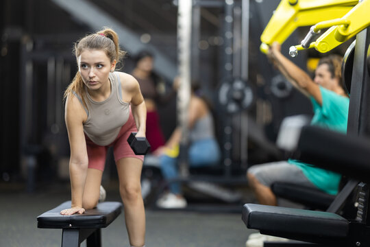 Young woman doing exercises with a dumbbell for back muscles. European woman performs a dumbbell row near a bench in the gym against the background of training people