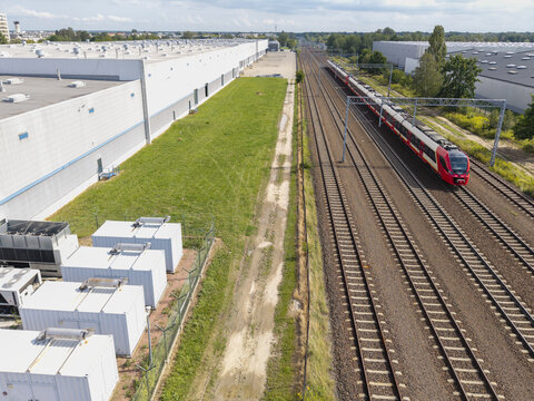 Aerial view of power generator units providing energy backup for an AI data center, situated next to railway tracks with a passing train and large industrial warehouses in Poland.