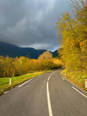 Naklejka premium Winding country road runs through glowing autumn trees while dark clouds gather above the hills. Striking contrast between golden foliage and the moody sky. Journey through countryside in fall season.