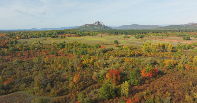 AERIAL: Colourful autumn landscape with a rocky peak rising beyond meadows, shrubs, and mixed woodland. Patchwork fields, turning leaves and distant hills create a broad scenic panorama in Croatia.