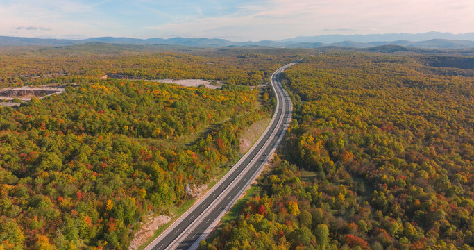 AERIAL: Winding motorway stretching across a vast autumn forest. Smooth asphalt road snakes through colorful hills and woodland. Travelling through rolling landscape in warm shades of fall season.