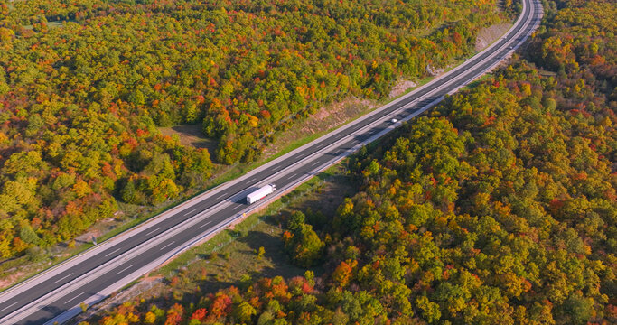 AERIAL: White truck drives along motorway cutting through sunlit autumn woodland. Light traffic moves along modern asphalt route bordered by vivid foliage, rocky slopes and scenic rolling countryside