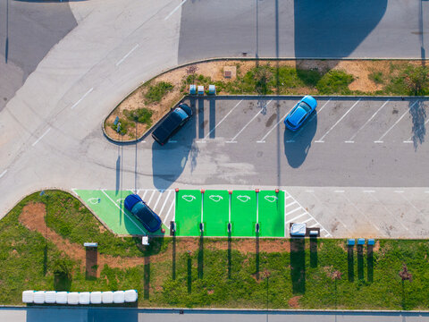 AERIAL TOP DOWN: Cars are parked beside green electric vehicle charging spaces at a roadside facility. View of modern transport infrastructure with green marked spaces, charging units and parked cars.