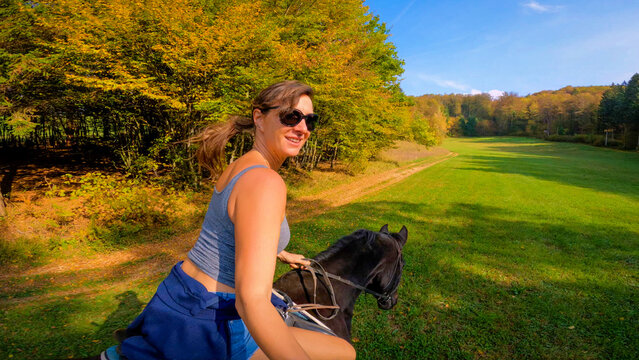 SELFIE: Smiling young woman riding a dark brown horse along a green meadow while colorful autumn foliage decorates the surrounding forest under a clear blue sky. Joyful equestrian moment in Slovenia.