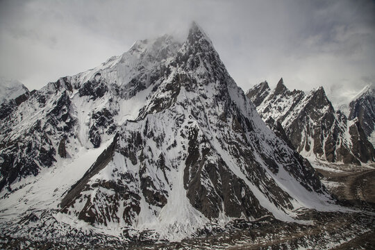 Aerial view of Mitre Peak, a majestic snow-covered mountain, rising above the rugged Baltoro Glacier with dramatic rocky ridges and glacial ice under a cloudy sky, Skardu, Pakistan.