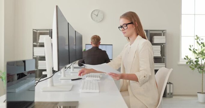 Businesswoman analyst reviewing spreadsheet data on monitors. Professional in a bright office reviews finance analytics, typing notes to ensure accuracy. Clear business data analysis concept.
