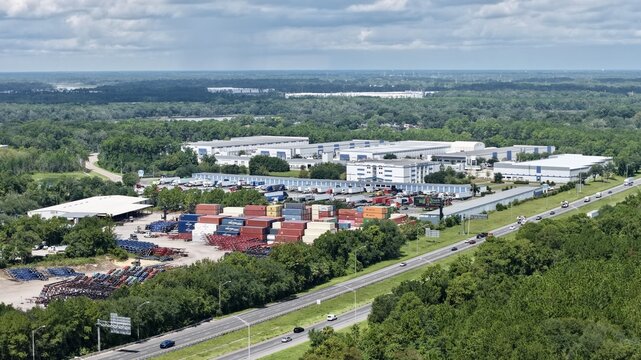 Aerial view of a logistics facility featuring rows of shipping containers, large warehouse buildings, and a nearby highway surrounded by dense green forest in Jacksonville, Florida, United States.
