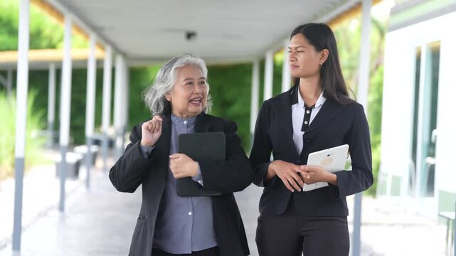 Businesswoman and colleague conversation while walking corridor with tablet and folder, mentoring during professional meeting, senior and young professionals discuss project strategy near building