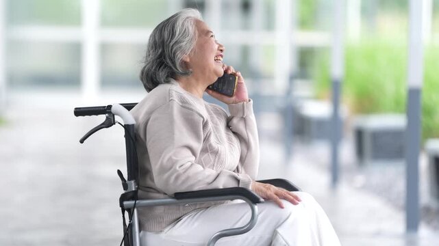 Senior woman in wheelchair using smartphone for phone call, smiling outdoor with caregiver support, elderly asian patient healthcare communication on patio, active aging independence living
